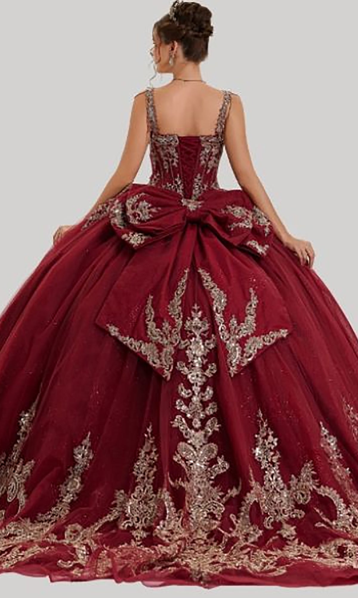 A woman is shown from behind against a plain background wearing the Quinceanera Dress 26407 by Stella Couture—an elaborate burgundy ball gown featuring silver embroidery, a dramatic bow at the back, and a voluminous glitter tulle skirt adorned with embroidered appliques.